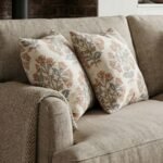 Premium designer cushion covers in beige cotton fabric featuring hand-block style rust floral motifs with sage green leaves, styled for luxury living room and bedroom interiors.