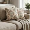 Premium designer cushion covers in beige cotton fabric featuring hand-block style rust floral motifs with sage green leaves, styled for luxury living room and bedroom interiors.