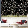 Daisy Bennett mural wallpaper by RUBY featuring delicate floral trees with blossoms and birds on a black background, styled in a modern luxury living room with a white sofa and soft decor