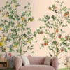 Elegant botanical mural wallpaper featuring hand-painted citrus trees with oranges, lemons, and delicate birds on a soft peach background, styled in a modern living room with a green velvet sofa and natural wood furniture.