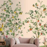 Elegant botanical mural wallpaper featuring hand-painted citrus trees with oranges, lemons, and delicate birds on a soft peach background, styled in a modern living room with a green velvet sofa and natural wood furniture.